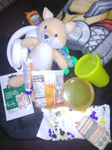Folded blanket, fox stuffed animal, white headphones, goldfish, gummies, baby wipes, tissue, monster truck, hand sanitizer, 1st aid mini kit, sunscreen, snack bowl with lid, and yellow sippy cup with reusable metal straw on black mesh bag