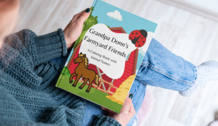 Woman wearing blue sweater and blue jeans holding "Grandpa Donn's farmyard friends" coloring book.