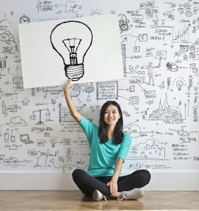 woman holding lightbulb with drawing on a white wall behind her; she is seating on a wood floor