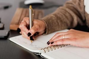 woman wearing sweater with black nails writing in notebook with gold /silver pen. background is an office with keyboard.